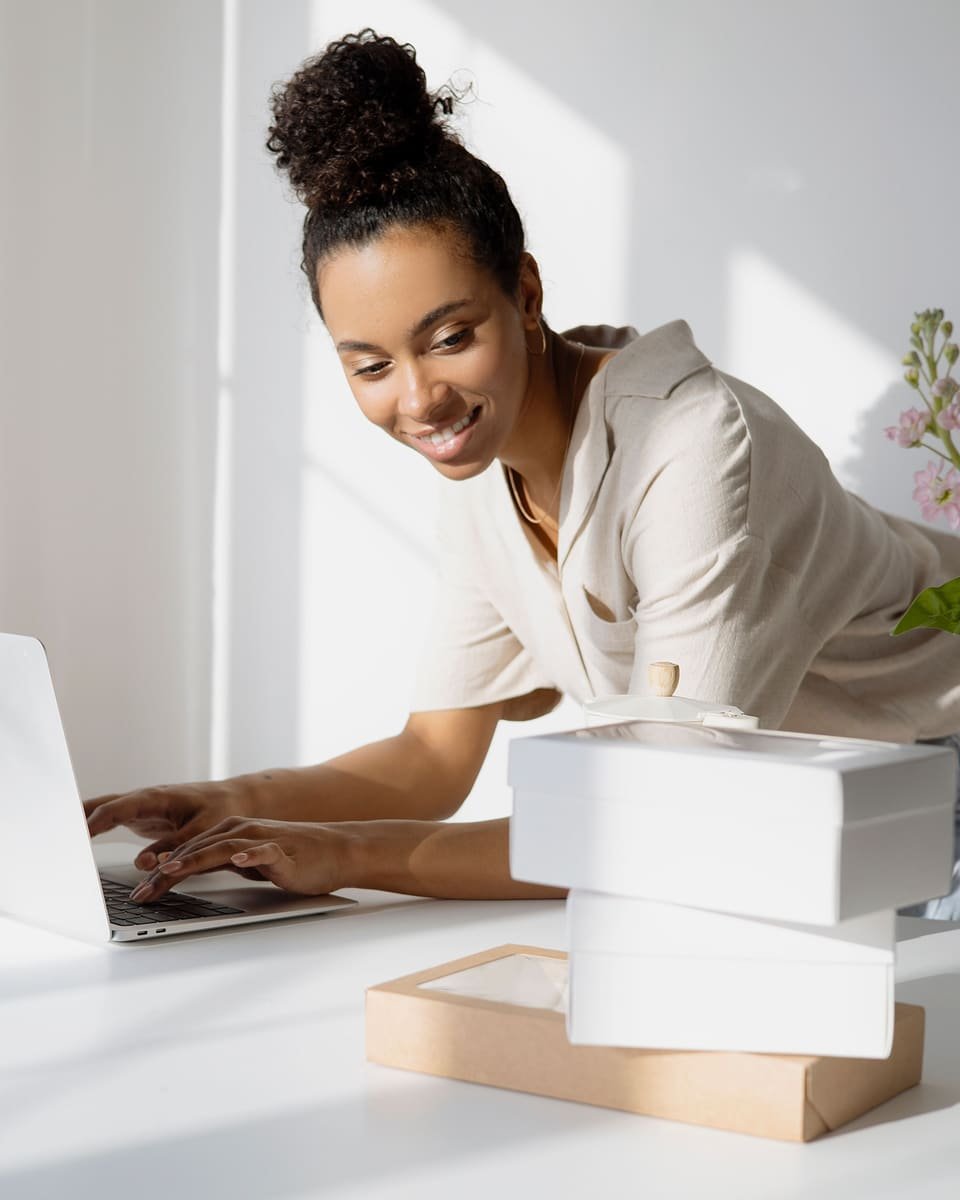 Empreendedora sorri enquanto digita no notebook, ao lado de caixas de envio e de um vaso de flores